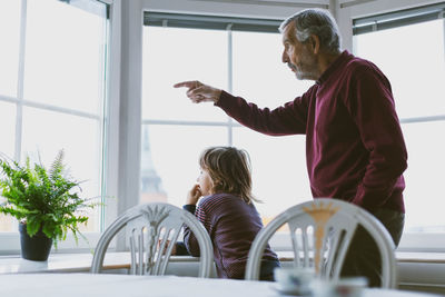 Senior man showing something to great grandson through window at home