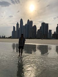 Man standing on sea by buildings against sky during sunset