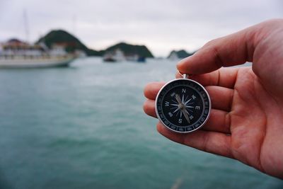 Close-up of hand holding navigational compass over sea against sky