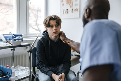 Male doctor checking neck of teenage patient while sitting in medical examination room