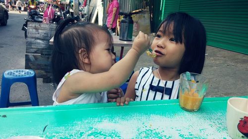 Cute girl feeding sister at sidewalk cafe in city