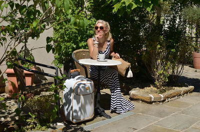 Woman in elegant dress sitting in mediterranean yard with elegant suitcase