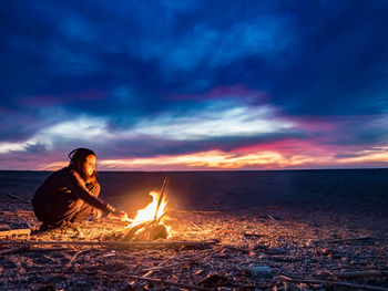 Woman crouching by campfire during sunset