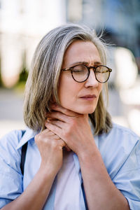 Sad senior woman wearing glasses holding her sore throat outdoors across the city.