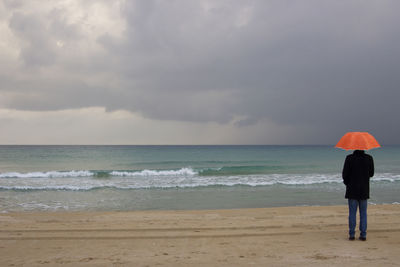Rear view of man with umbrella standing at sea shore against cloudy sky