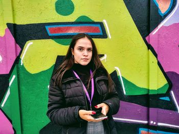 Portrait of young woman holding mobile phone against colorful graffiti wall