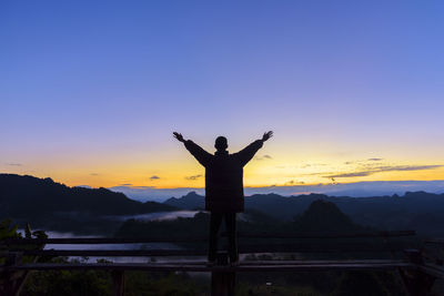 Rear view of silhouette man with arms outstretched standing on railing against sky during sunset