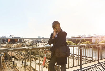 Smiling young man talking on smart phone while standing with electric push scooter at bridge in city against sky