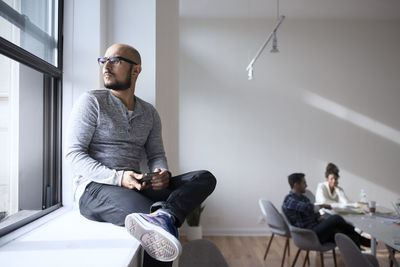 Thoughtful businessman sitting on window with colleagues in background