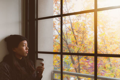 Young woman looking through window while holding disposable cup