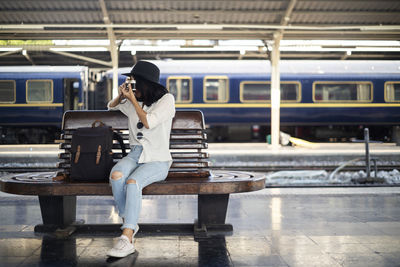 Full length of woman holding camera sitting at railroad station platform