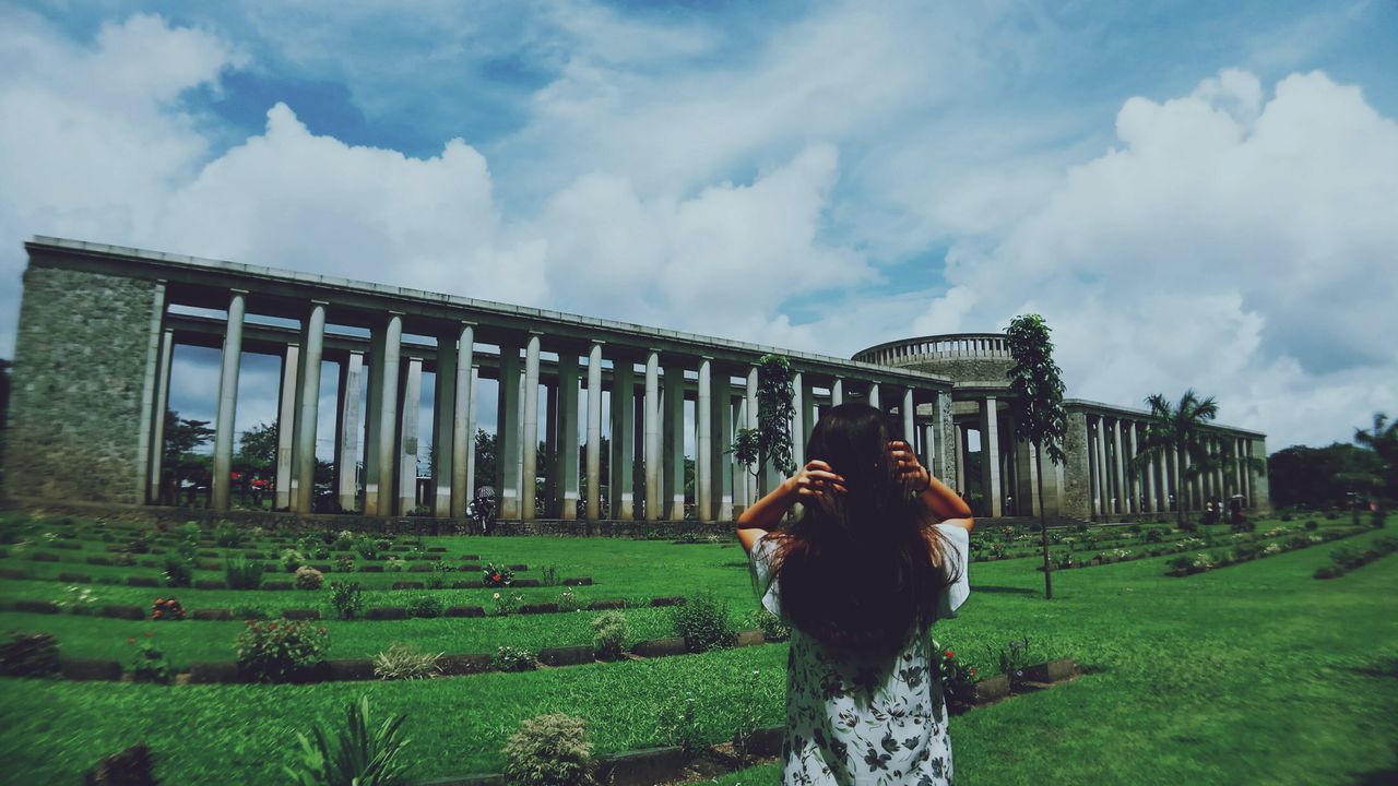 cloud - sky, real people, one person, architecture, sky, lifestyles, leisure activity, standing, built structure, rear view, nature, grass, three quarter length, women, adult, building exterior, day, plant, field, outdoors, hairstyle