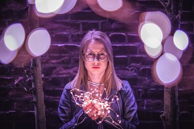 Portrait of young woman holding illuminated string lights at night