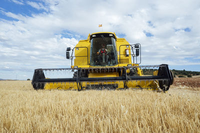 Combine harvester with metal reel collecting dried wheat in agricultural plantation against cloudy sky in rural area with green trees