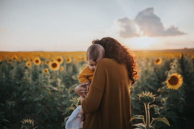 Mother carrying toddler daughter in sunflower farm against sky