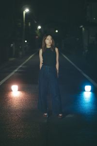 Full length portrait of young woman standing on illuminated street at night