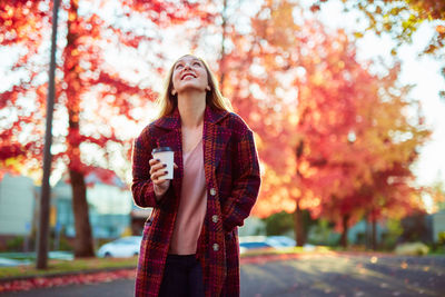 Young woman holding coffee while standing against autumn trees