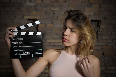 Young woman holding film slate against brick wall