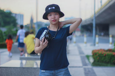 Portrait of young woman holding camera while standing on footpath in city
