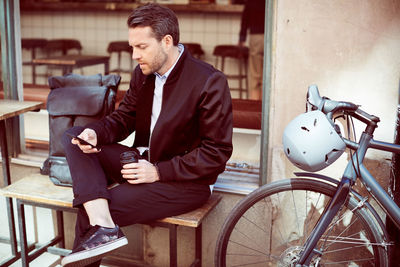 Businessman using mobile phone while sitting on bench by bicycle in city