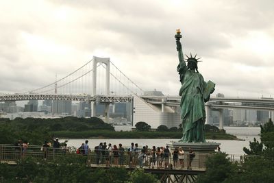 Statue of suspension bridge in city against cloudy sky