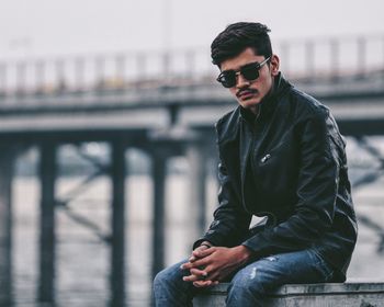 Portrait of young man sitting on railing in city against sky