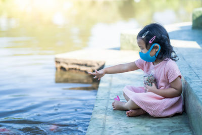 Cute girl wearing protective face mask feeding carp koi fishes at pond.