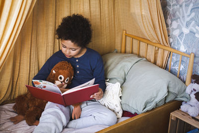 Boy with stuff toy reading story book while sitting on bed at home