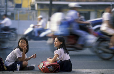 Blurred motion of people by schoolgirls sitting on street