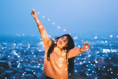 Cheerful woman holding illuminated string light against clear sky at dusk