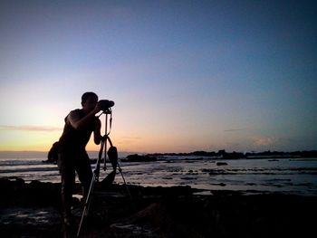 Man photographing at beach against clear sky during sunset