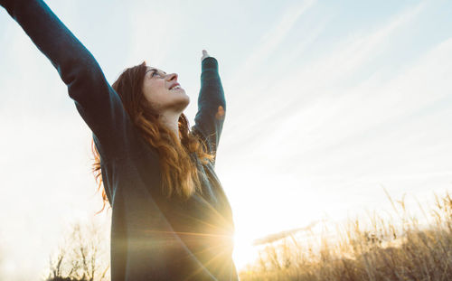 Smiling woman with arms raised standing against sky