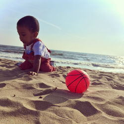 Boy kneeling by ball on sand at beach