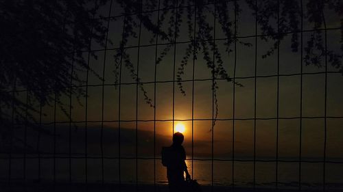 Silhouette man standing by fence against sky during sunset