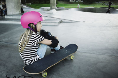 Thoughtful girl wearing pink helmet sitting at skateboard park