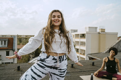 Portrait of smiling young woman practicing yoga by friend on rooftop