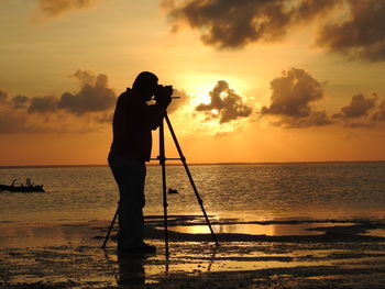 Silhouette man photographing at beach using camera against sky during sunset