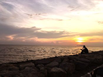 Silhouette man sitting on rocks by sea against sky during sunset