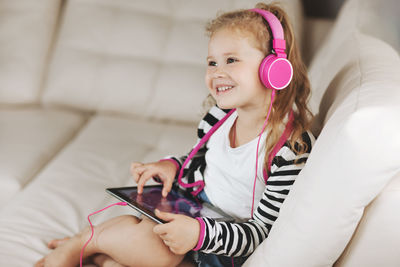 A little girl in pink headphones is sitting on the couch playing on a tablet and listening to music