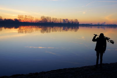 Rear view of silhouette man holding gun by lake against sky during sunset