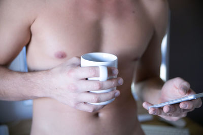 Midsection of shirtless man with coffee mug using smart phone at home