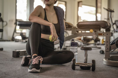 Low section of woman holding fruit sitting in gym