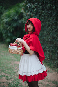 Woman in little red riding hood costume holding basket while standing against building