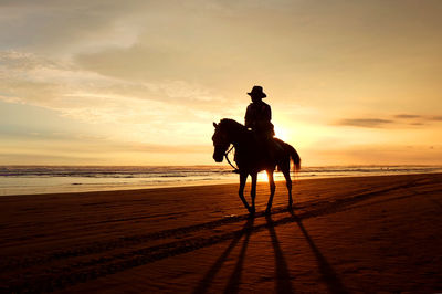 Silhouette people riding horse on beach against sunset sky