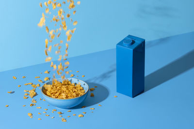 Pouring cereals in a bowl on blue table. breakfast table, cornflakes cereals in bowl and a milk box