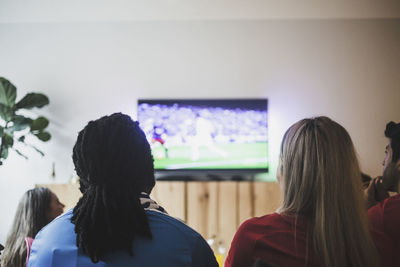 Rear view of friends watching soccer match while sitting at living room