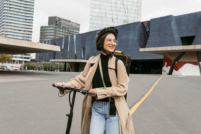 Smiling woman wearing cycling helmet looking away while walking with electric push scooter at city