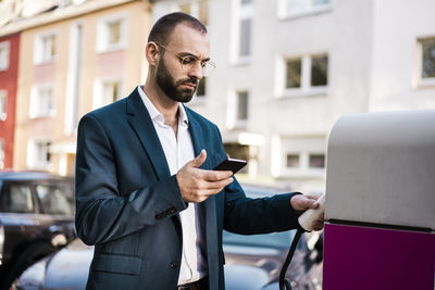 Businessman holding mobile phone at electric vehicle charging station