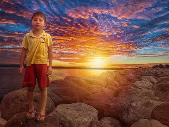 Boy standing on rock against sky during sunset