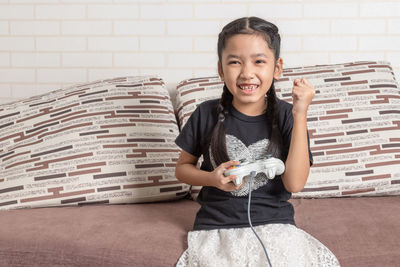 The little asian girl playing video games with a joystick and sitting on the sofa with happiness.